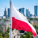 Na imagem destacada contem a bandeira da polônia nas cores vermelha e branca com a cidade de fundo
